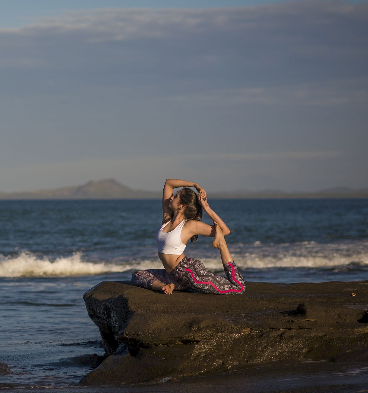 A person practices yoga on a rock by the ocean shore under a clear sky, with mountains visible in the distance and waves approaching the shore.