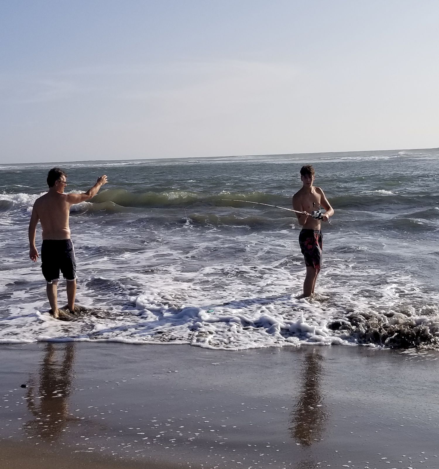 Two people standing in the shallow water of a beach, with waves washing up around them, both wearing shorts against the backdrop of the ocean.