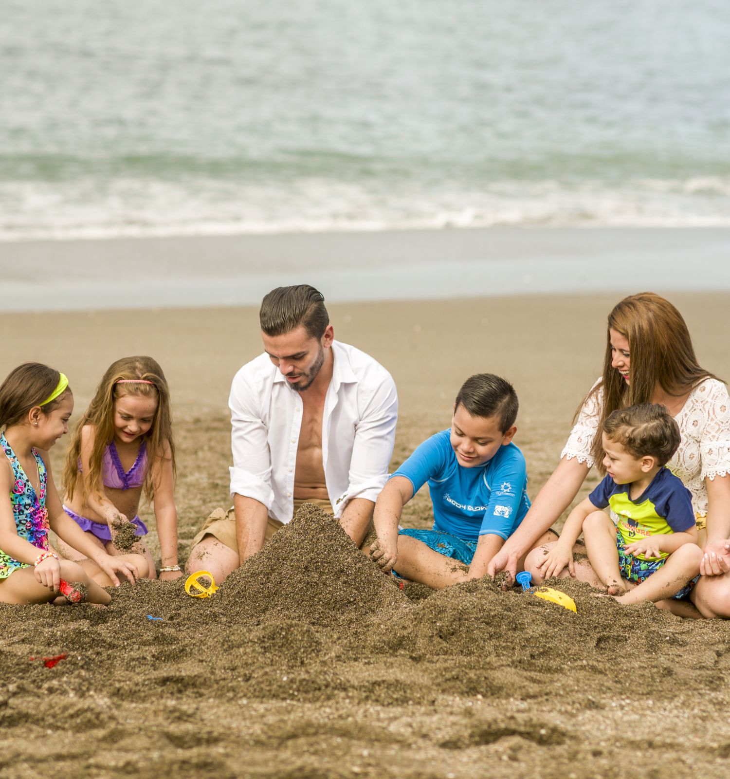 A family with three children is sitting on the beach, building a sandcastle together, with the ocean in the background.