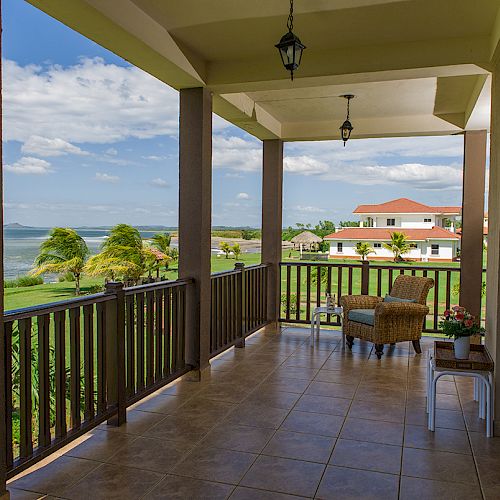 A scenic porch with wooden railings, wicker chairs, and a view of greenery and distant water under a cloudy sky.