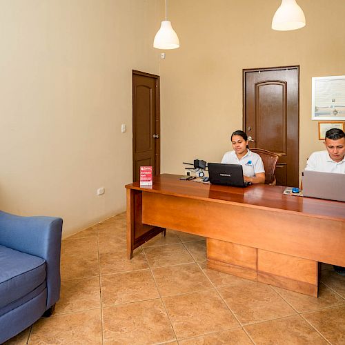 The image shows two people working at a desk on laptops in an office with a blue chair, beige walls, and overhead lighting.