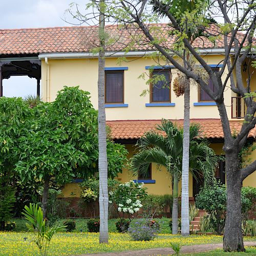 A two-story yellow house with a red-tiled roof, surrounded by lush greenery, flowers, and trees, is shown in the image.