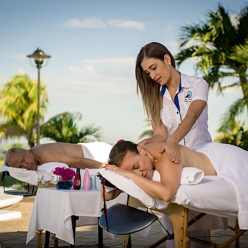 Two people are receiving outdoor massages near a pool, surrounded by tropical plants and blue skies, with a massage therapist attending to one person.