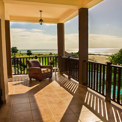 A patio with a wicker chair, overlooking a lush garden and a serene beach with the ocean in the background. The sky is sunny and clear.