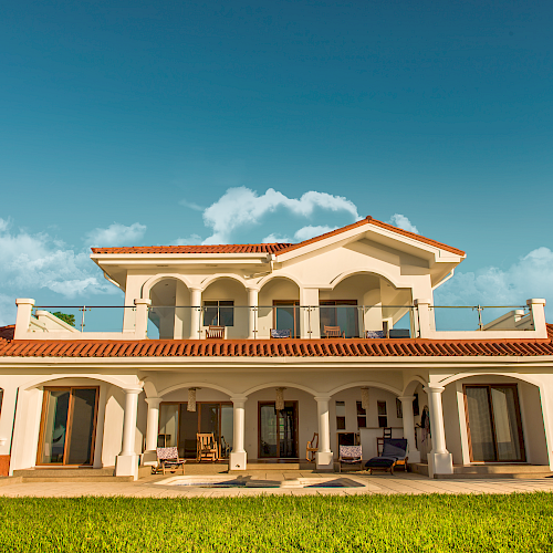 A large, two-story house with a red-tiled roof, arched windows and doors, and a spacious lawn under a partly cloudy sky ends the sentence.