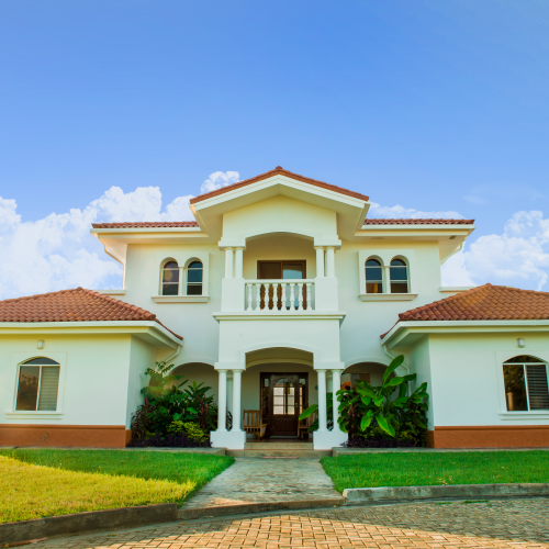 A large, two-story house with a red-tiled roof, white exterior, and landscaped lawn under a clear blue sky with clouds.