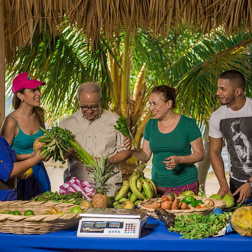 A group of people are gathered at a market stall with fresh produce, including greens, bananas, and other vegetables, near a beach.