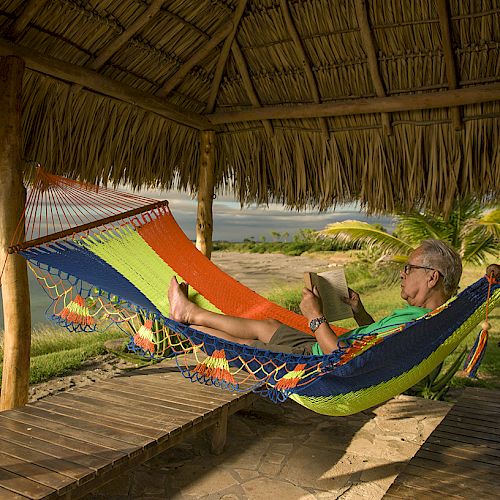 A person relaxes in a colorful hammock under a thatched gazebo, reading a book with an ocean view in the background, surrounded by lush greenery.