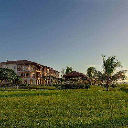 A large building with balconies overlooks a lush, green lawn dotted with palm trees and a gazebo, near a body of water, under a clear sky.