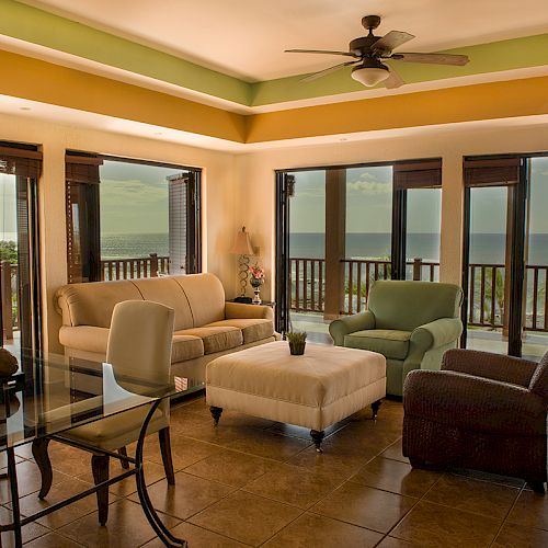 A spacious living room with multiple seating arrangements, large windows, ocean view, wooden flooring, and a ceiling fan is pictured in this image.