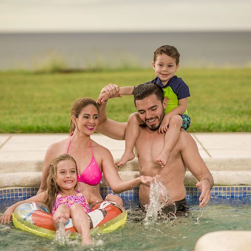 A family of four enjoys a pool, with the father carrying the son on his shoulders, the mother and daughter in front, smiling and splashing water, ending the sentence.