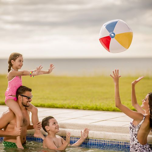 A family enjoys time in a pool; a man holds a girl on his shoulders while a woman throws a beach ball, and a boy reaches for it.