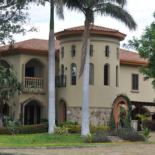 A two-story Mediterranean-style house with a round turret, surrounded by palm trees and greenery.