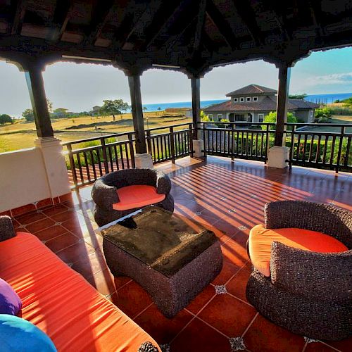 A spacious, covered outdoor patio with wicker furniture and colorful cushions overlooking a scenic landscape with a house and water in the distance.