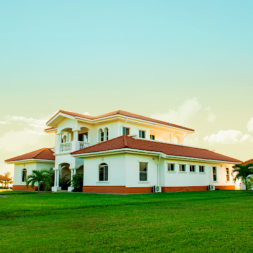 The image shows a large, modern house with a red-tiled roof and white walls, surrounded by a spacious green lawn and a clear blue sky.