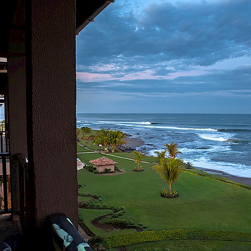 A view from a balcony overlooks a manicured lawn with palm trees, a gazebo, and a coastline with ocean waves under a partly cloudy sky.