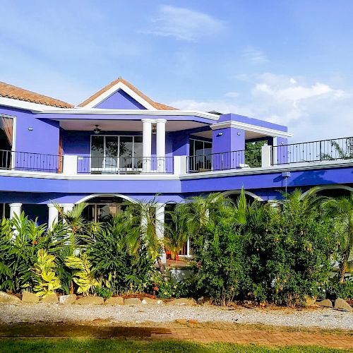 A vibrant blue two-story house with a balcony, surrounded by lush greenery and a well-maintained garden, on a sunny day.