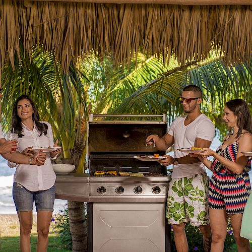 A group of friends enjoying a barbecue under a thatched roof by the beach, with palm trees in the background.