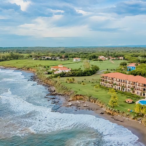 A coastal resort featuring a swimming pool and buildings with red roofs, lush green landscape, and the ocean waves crashing onto a rocky shore.