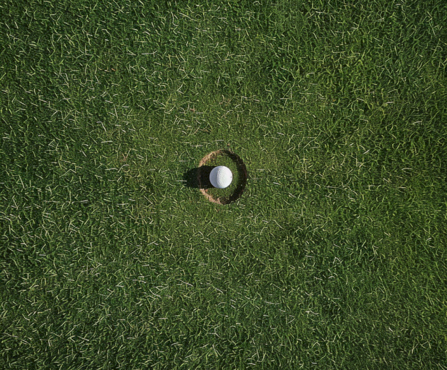 A white golf ball is in close proximity to a hole on a green golf course, casting a shadow. The image is taken from above.