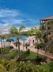 A tropical resort by the sea, with a pool, green lawns, palm trees, and a building on the right side. Blue sky and ocean in the background end the sentence.