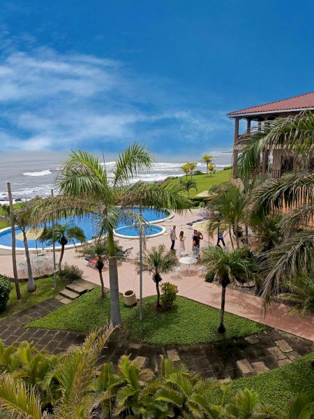 A tropical resort by the sea, with a pool, green lawns, palm trees, and a building on the right side. Blue sky and ocean in the background end the sentence.