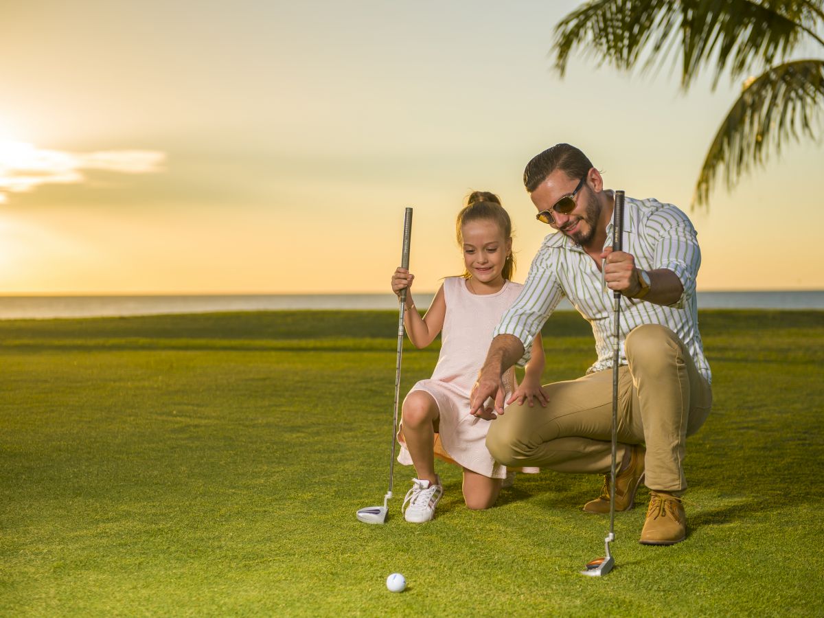 A man kneels on the grass, teaching a young girl how to putt a golf ball, with a sunset and palm tree in the background.