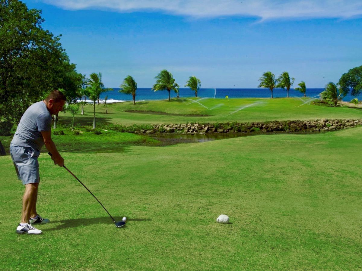 A person is preparing to hit a golf ball on a lush green golf course overlooking the ocean, with palm trees lining the horizon.