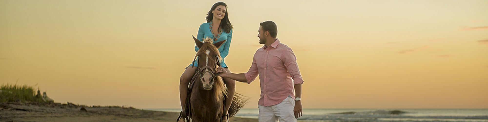 A woman sits on a horse while a man walks beside them on a beach at sunset. They are enjoying a peaceful moment by the ocean.