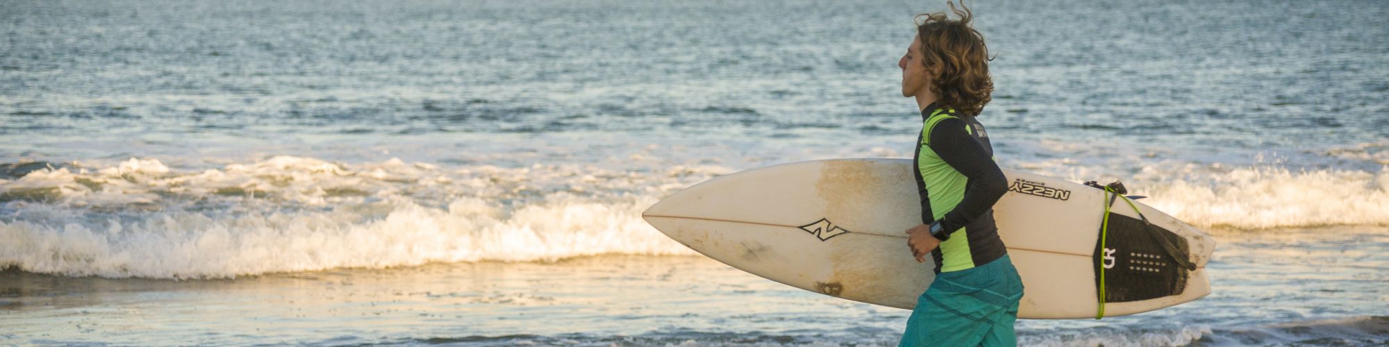 A person in a wetsuit is holding a surfboard and jogging towards the ocean on a beach, with waves in the background and a distant landmass.