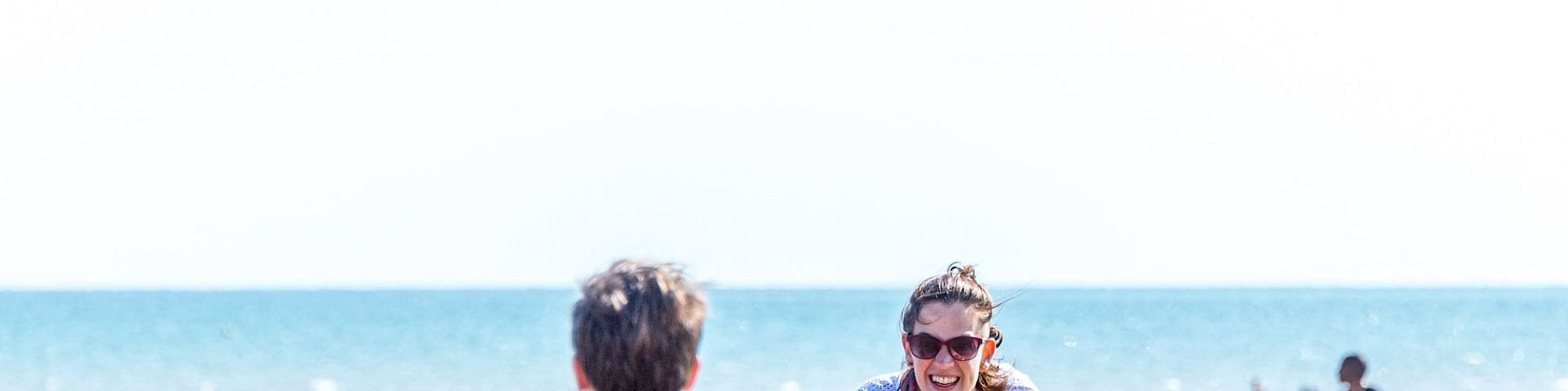 A woman and a boy are playing on a sunny beach, with other people and the sea in the background. The woman is smiling and leaning toward the boy.