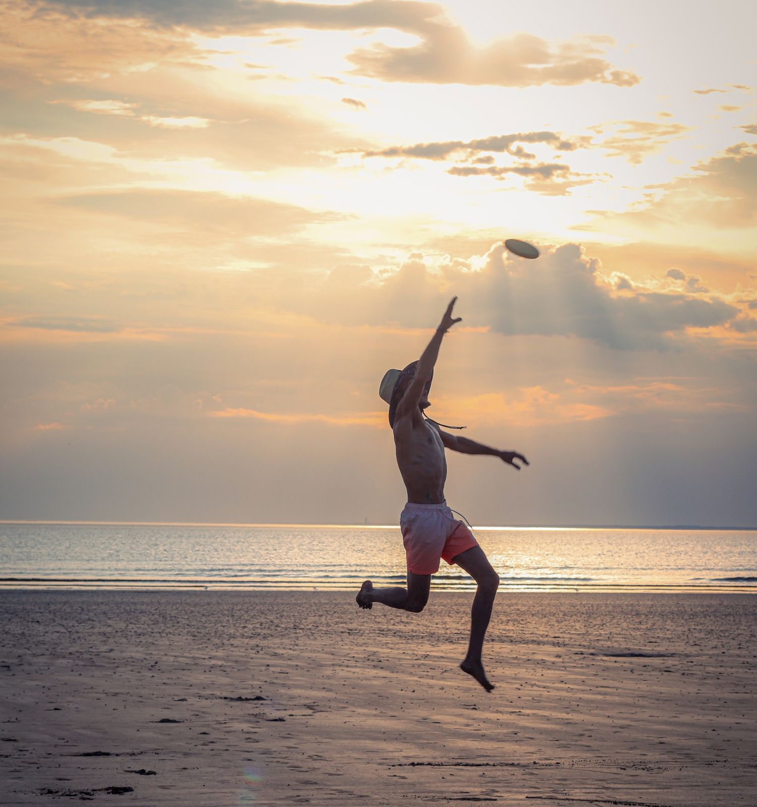 A person is jumping and reaching toward the sky on a beach during sunset, with the sun setting over the ocean.