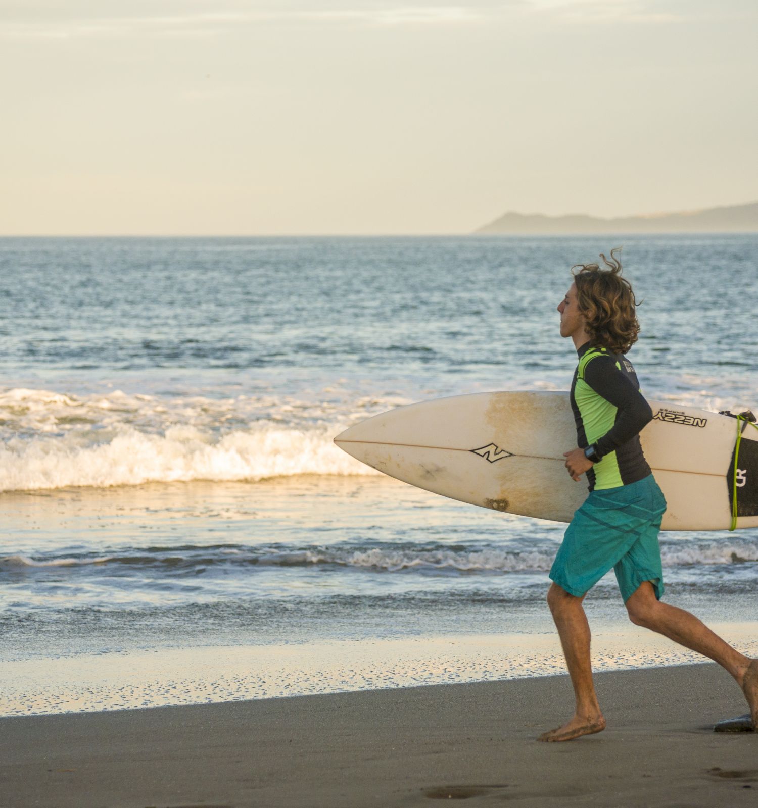 A person is running along the beach carrying a surfboard, with waves and distant mountains in the background.