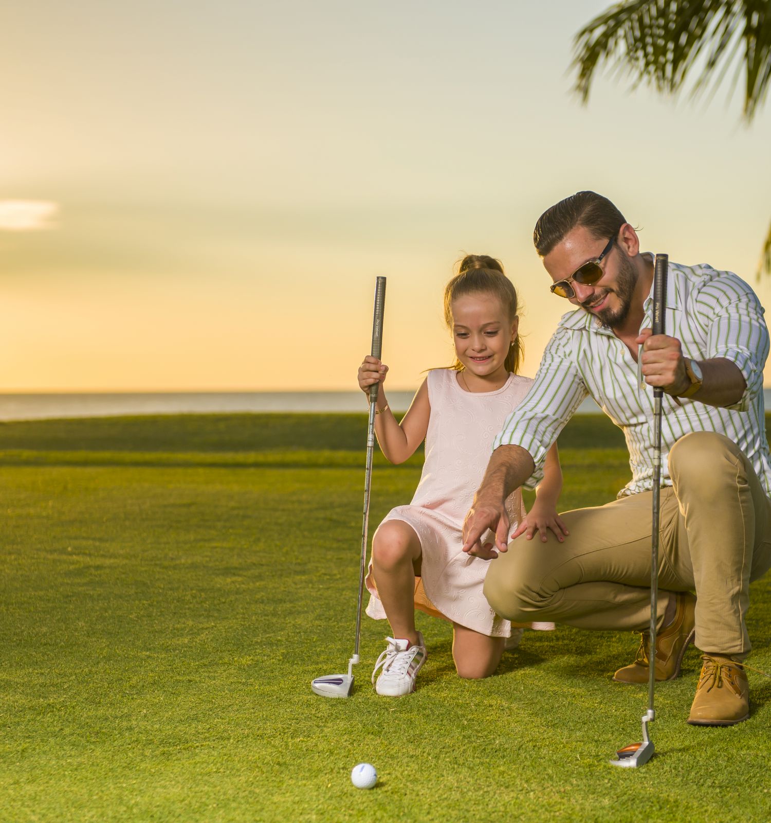 A man and a child, both holding golf clubs, are kneeling on a golf course with a palm tree and sunset in the background.