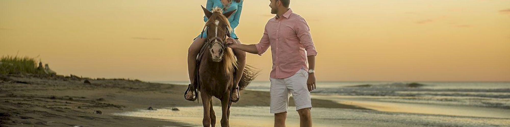 A woman rides a horse on the beach at sunset while a man walks beside them, holding the horse's reins, creating a serene seaside scene.