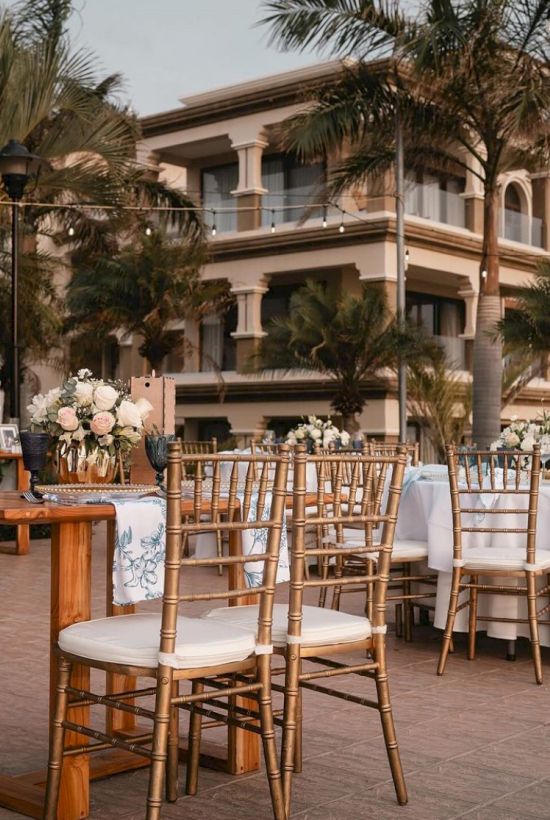 An outdoor setup with chiavari chairs and decorated tables, surrounded by palm trees and a modern building in the background.