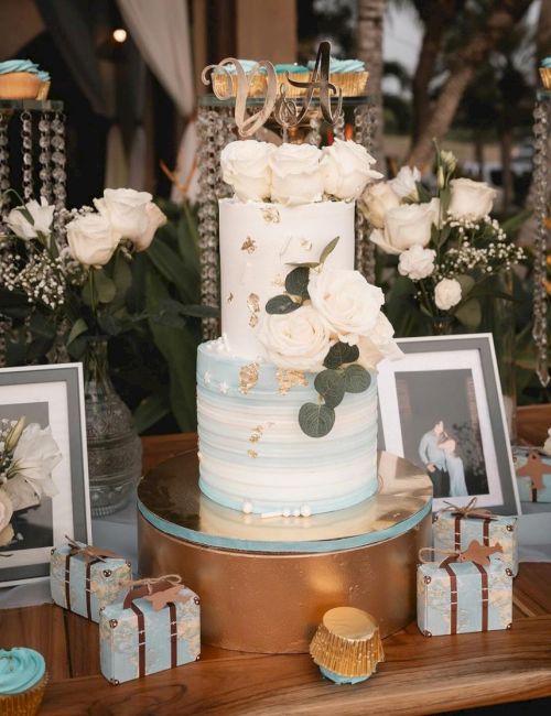 The image features a beautifully decorated cake with white roses, surrounded by flowers, framed photos, small gift boxes, and cupcakes on a wooden table.