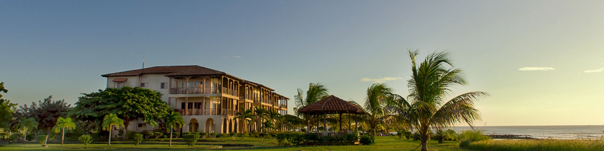 A large building surrounded by lush green grass, palm trees, and a small gazebo, with a serene ocean view under a clear sky.