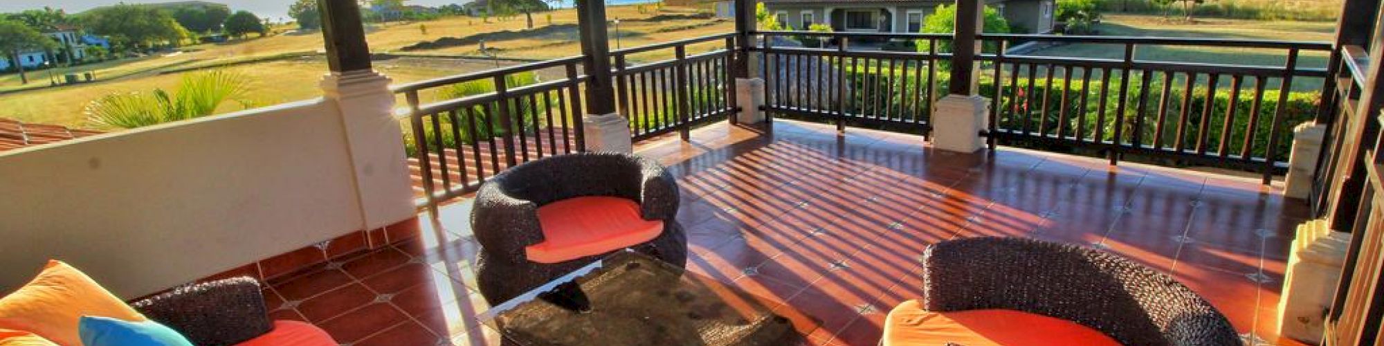 An outdoor covered patio with wicker furniture, colorful cushions, and a scenic view of a house, fields, and the ocean in the background.