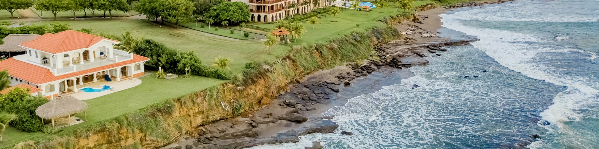 An aerial view of a coastal area with a large house, a pool, and a building, all surrounded by greenery, near a rocky shoreline and ocean waves ending the sentence.