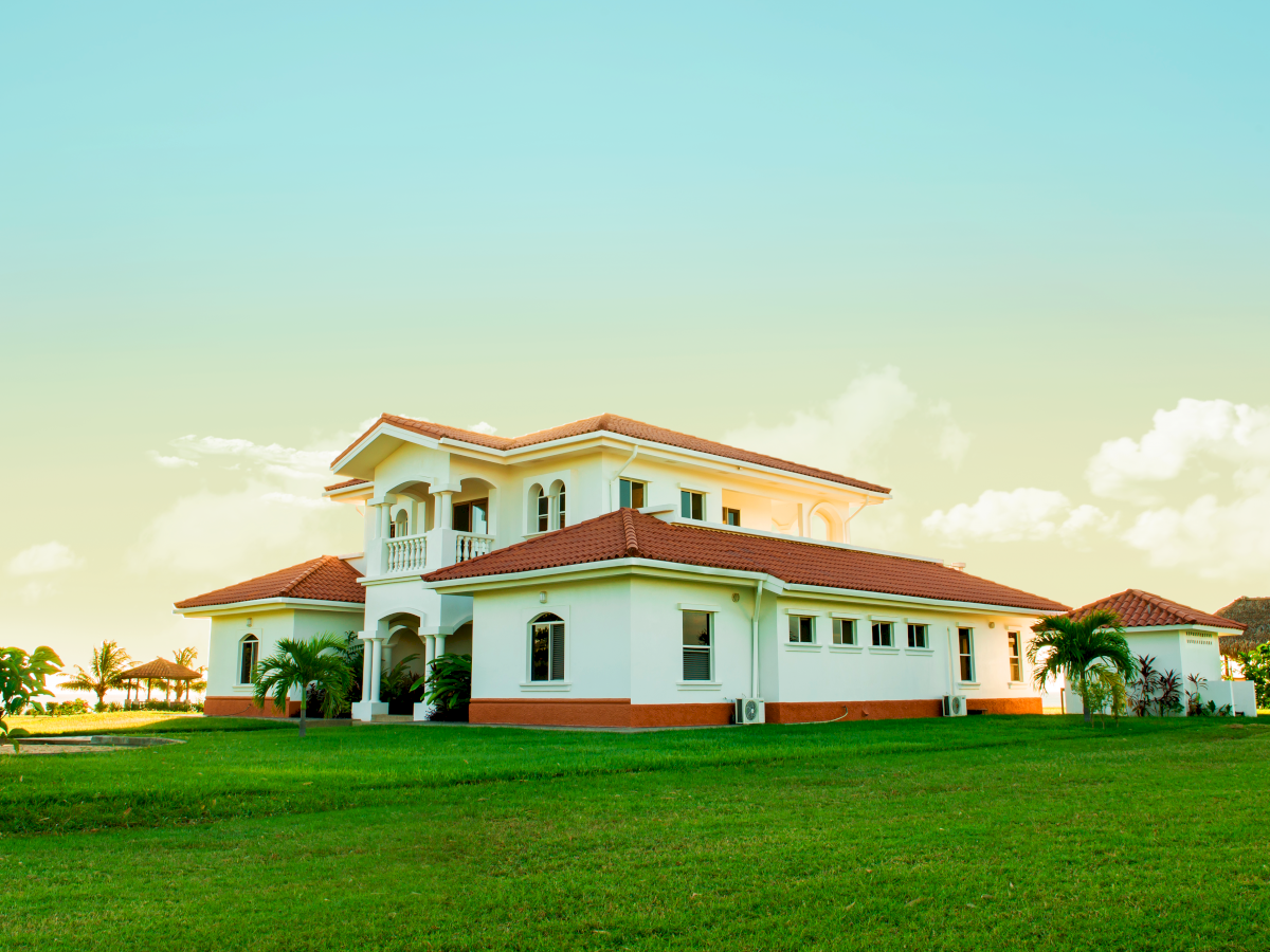 A large white house with a red roof sits on a well-manicured lawn, with palm trees around and a clear, bright sky in the background.