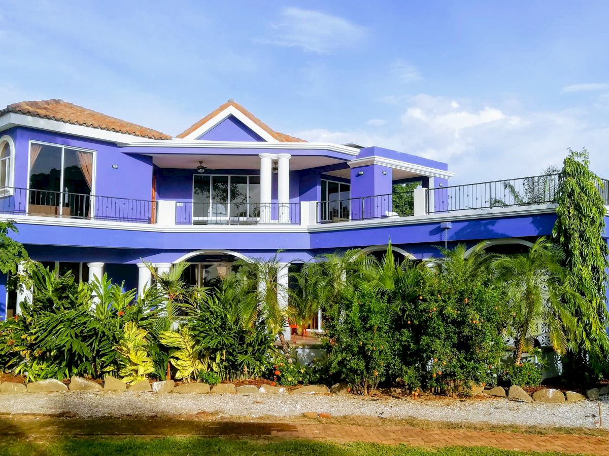 A two-story blue house with a tiled roof, surrounded by greenery, featuring large windows and balconies under a clear sky.