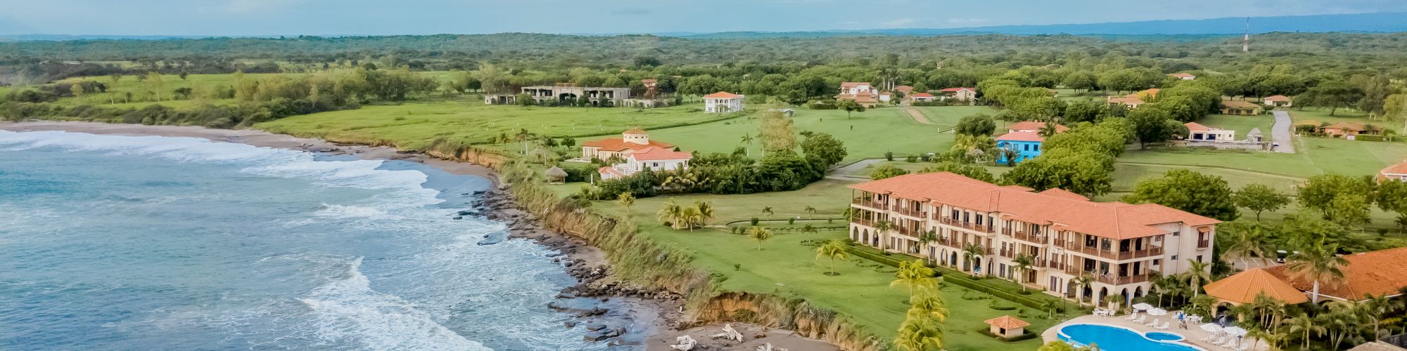 This image shows a coastal landscape with waves hitting the shore, a sandy beach, a large building with a pool, greenery, and scattered small buildings.