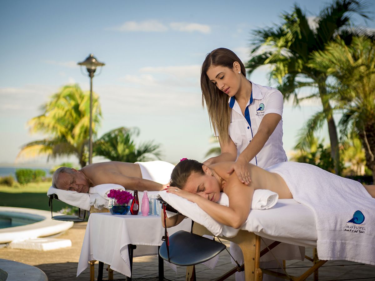 Two people are receiving massages by a poolside, surrounded by palm trees and tropical scenery, while a masseuse tends to one of them.