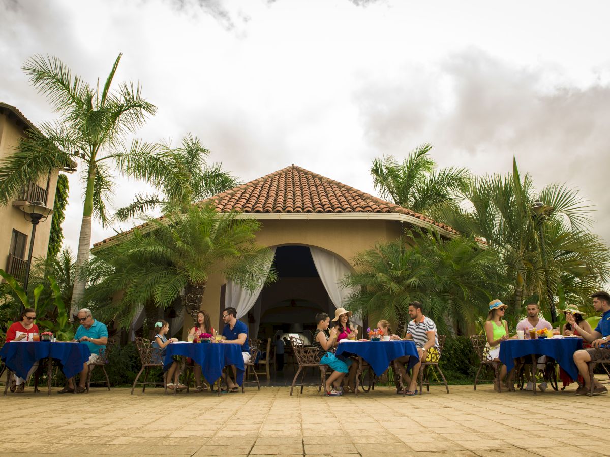 People are sitting at tables with blue tablecloths outside a building with a tiled roof, surrounded by palm trees on a cloudy day.