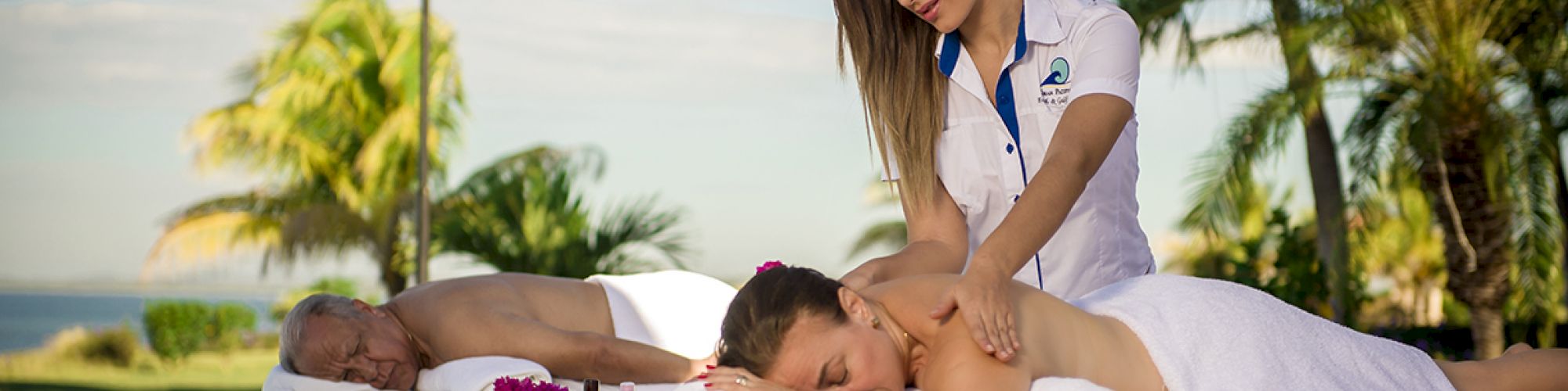 Two people are receiving outdoor massages near a pool, surrounded by palm trees. A therapist is massaging one client, while the other relaxes.