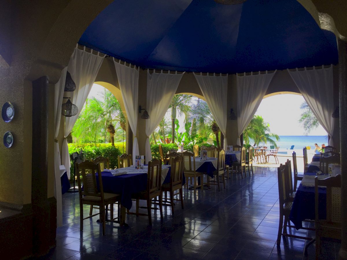 The image shows an open-air restaurant with wooden tables and chairs, tropical plants, and a view of the beach and ocean beyond.