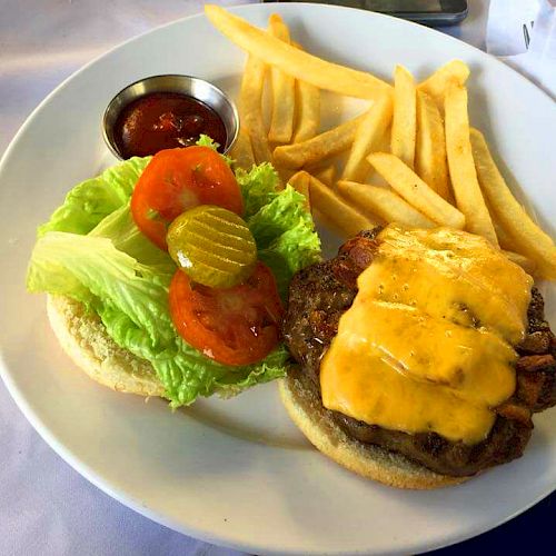 This image shows a cheeseburger with lettuce, tomato, and pickles, along with a side of fries and a small container of ketchup on a white plate.