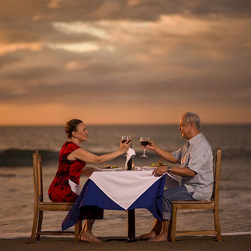 A couple is having a romantic dinner on the beach at sunset, toasting with wine glasses, with a table set with a white and blue tablecloth.