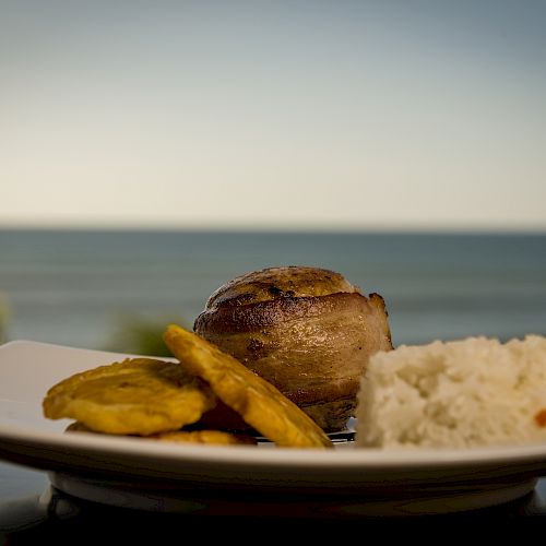 A dish with rice, plantains, and a piece of meat, photographed in front of a sea view, served on a white plate.